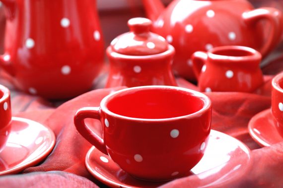 A cup of tea alongside teapots, milk jug and sugar bowl, all red china with white spotted pattern