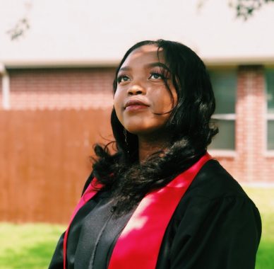 University graduate looking upwards in black graduating gown with red trim, standing in front of building and grass