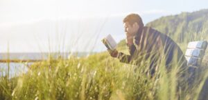 Man in coastal landscape with grass sitting on bench, reading book